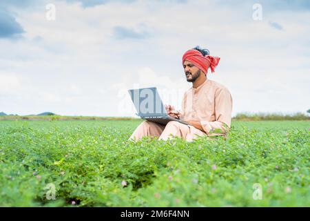 Young indian farmer busy wrking on laptop while sitting at agricultural farmland - concept of technology, digital communication and modern farming. Stock Photo