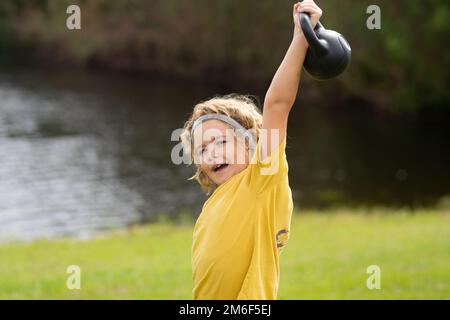 Kid lifting the kettlebell in park outside. Kid boy working out with dumbbells. Sport and kids training. Stock Photo