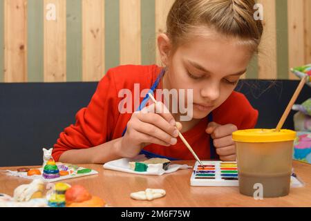 A girl dips a brush in paint of the desired color while painting a craft from salt dough Stock Photo