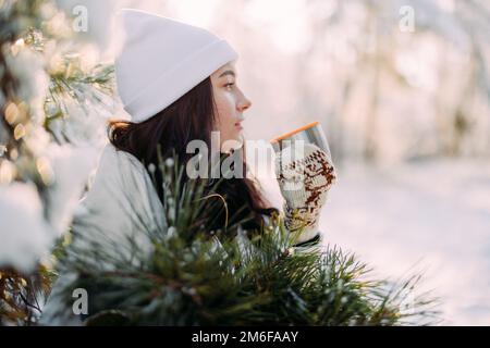 Happy young woman drinks hot tea from thermos cup during walk in winter forest with bouquet from pine branches in her hands. Stock Photo