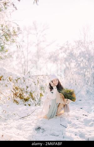 Happy young woman sits and smiles in forest among snow covered trees in sunny winter day with bouquet from pine branches in her hands. Stock Photo