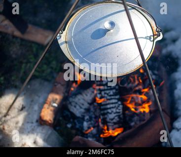 Black large tourist pot hangs over burning flame of campfire in the forest.  Cooking dinner on