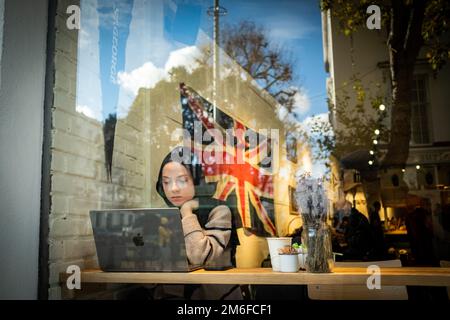 London- December 2022:  A woman in a cafe on Kings Road Chelsea working on a laptop with a British union flag behind Stock Photo