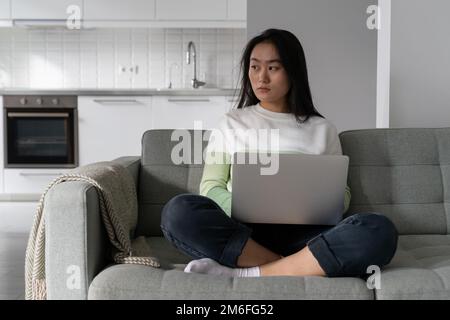 Young thoughtful Asian woman holding laptop on laps looking in distance waiting for important email Stock Photo