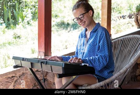 Young woman in glasses playing electric piano on terrace. Stock Photo