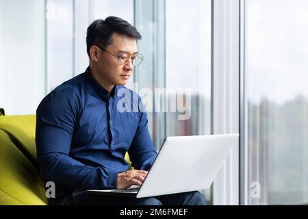 Serious and focused engineer developer programmer writing code using laptop at work to create programs and software, asian man inside modern office near window working. Stock Photo