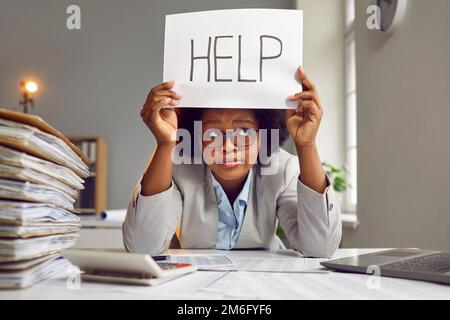 Tired, busy woman sitting at an office desk with paperwork and holding a HELP sign Stock Photo