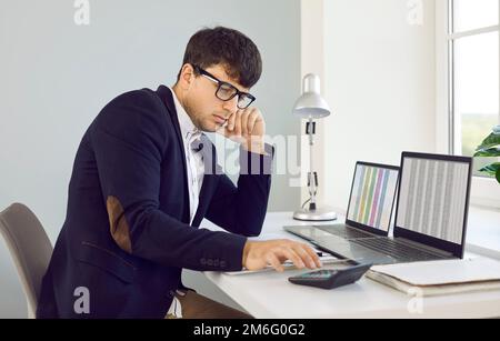 Accountant sitting at his office desk with laptops, looking at the calculator and thinking Stock Photo