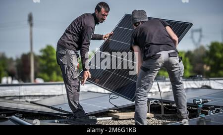 Male team engineers installing stand-alone solar photovoltaic panel system. Electricians mounting blue solar module on roof of company. Alternative en Stock Photo