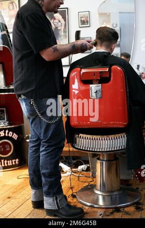 Barber is cutting young man hair. Back view of man getting short hair trimming at barber shop with a clipper machine. Stock Photo