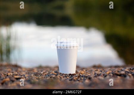 White paper cup of coffee or tea stands on the rocky shore of the lake Stock Photo