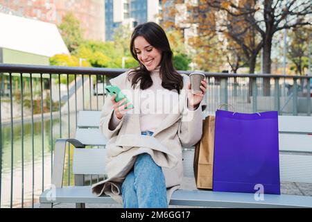 Happy young woman sitting on a bech using a smartphone to text messages after shopping on-line. Brunette female looking to the cellfone device sharing content on social media. High quality photo Stock Photo