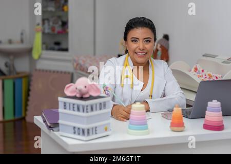portrait of a latina pediatrician doctor, sitting in her office making notes, while smiling looking at the camera. Stock Photo