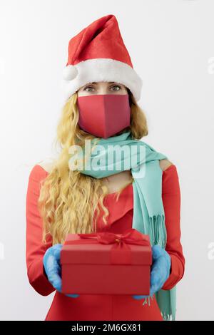 Lady Santa in a protective mask and gloves holds a box with a gift. Sterile gift. Protected christmas. Focus on the girl's face Stock Photo