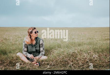 The girl is sitting cross-legged in the field, she is wearing dark glasses and is looking to the side into the distance Stock Photo