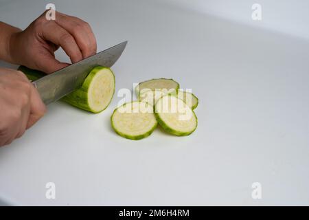 Female hand cutting zucchini. Hand is cutting green zucchini with a knife on a white background. Stock Photo