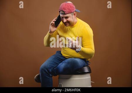 A man speaks expressively on the phone while sitting on a barrel in the studio. Chubby man talking on his smartphone emotionally Stock Photo