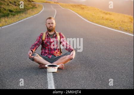 Positive bearded caucasian male tramp barefoot sitting on a country road at sunset with a passing charity donation hat Stock Photo