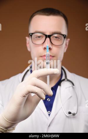 A young male doctor in glasses with a stethoscope around his neck and in a white coat is holding a plastic syringe with an injec Stock Photo