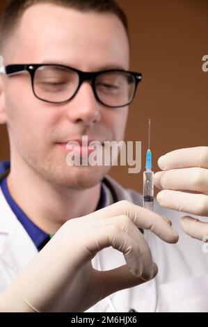 A young male doctor in glasses with a stethoscope around his neck and in a white coat is holding a plastic syringe with an injec Stock Photo