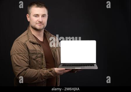 Attractive positive friendly Caucasian male holding laptop in hands presenting his cut out screen with copy space on black backg Stock Photo