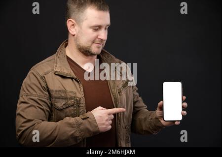 Caucasian attractive mature man in casual clothes holds a smartphone in his hands and points to the cut out phone screen with hi Stock Photo