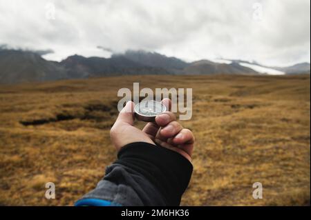 A man's hand of a traveler holds a magnetic compass against the background of a mountain landscape and low clouds on a sunny day Stock Photo