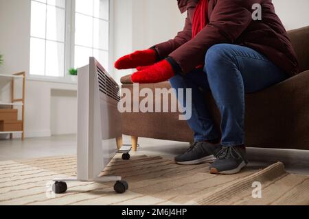 Young man is sitting at home on the sofa in warm jacket and knitted red mittens near heater, freezing from the cold. Stock Photo