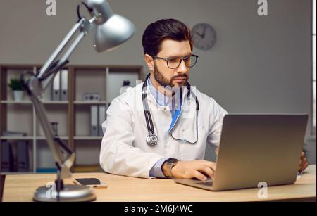 Young doctor working on a laptop computer while sitting at his desk at the hospital Stock Photo
