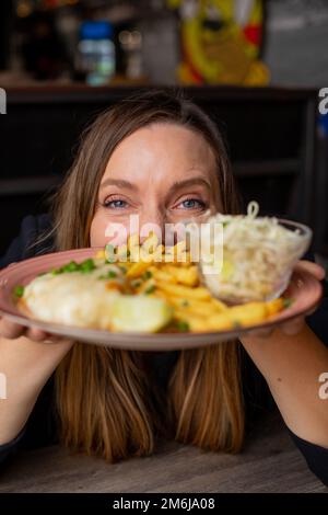 Cheerful woman gesturing hold in hands plate with fries, chicken fillet and vegetable salad. Serving of main dish and garnish closeup, selective focus Stock Photo