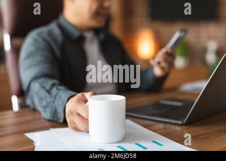 Smiling adult asian guy drinking cup of coffee, typing on smartphone at workplace with laptop in office interior Stock Photo