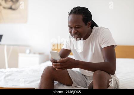 Smiling Black Man Messaging On Smartphone While Relaxing In Bedroom At Home Stock Photo