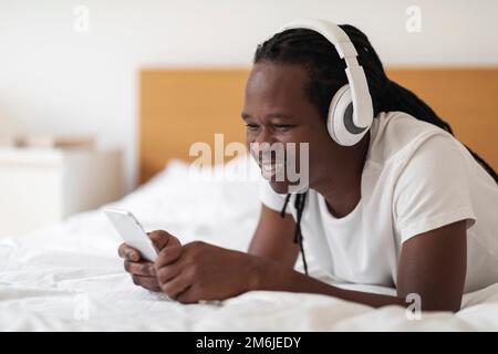 Smiling Young Black Man In Wireless Headphones Resting In Bed With Smartphone Stock Photo