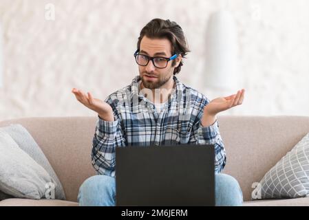 Surprised confused young caucasian guy blogger in glasses sits on sofa with computer, has video call Stock Photo