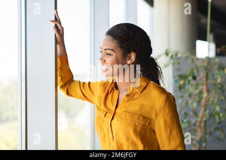 Successful african american businesswoman standing in modern coworking office and looking through window and smiling Stock Photo