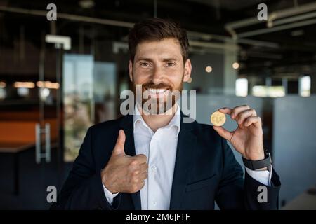 Happy middle aged businessman showing bitcoin and gesturing thumb up in modern office interior, free space Stock Photo