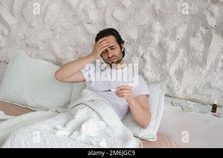 Unhappy sad young caucasian man suffers from headache and fever, look at thermometer, touch forehead with hand Stock Photo