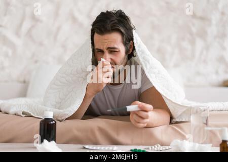 Despaired sad young caucasian sick male lies on bed under blanket, suffers from runny, blows his nose in napkin Stock Photo