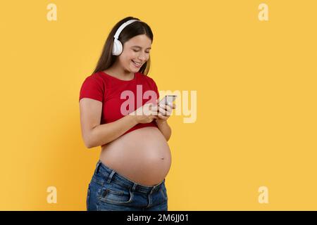 Modern Technologies. Happy Pregnant Woman In Wireless Headphones Listening Music On Smartphone Stock Photo