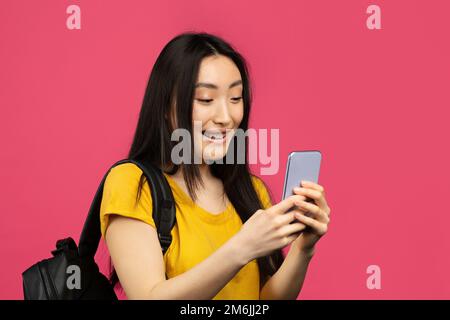 Happy asian female student using smartphone, texting online while standing with backpack on pink background, copy space Stock Photo