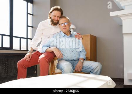 Father and son sit on the chair in the living room, both men look into the camera, an elderly man in glasses with diopters, a st Stock Photo
