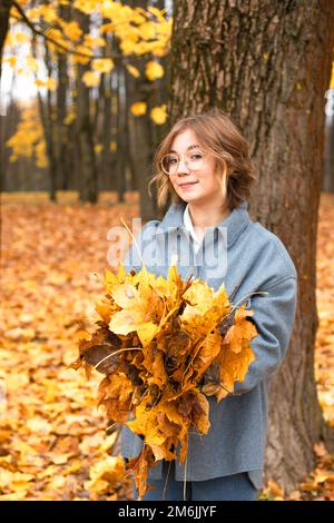 Portrait of young playful teenage girl standing among trees on yellow fallen leaves in park on sunny day in autumn. Stock Photo