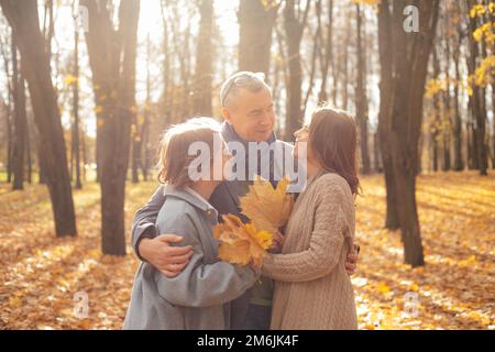 Portrait of loving family embracing, standing among trees in park forest, holding bunch of yellow fallen maple leaves. Stock Photo