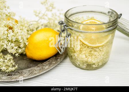 A jar of lemon and fresh elderberry inflorescences in syrup. Vintage metal plate on a white table. Stock Photo