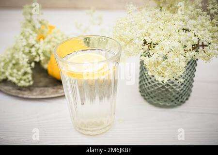 A faceted glass with a slice of lemon near a vase with fresh elderberry flowers on a white background. Stock Photo