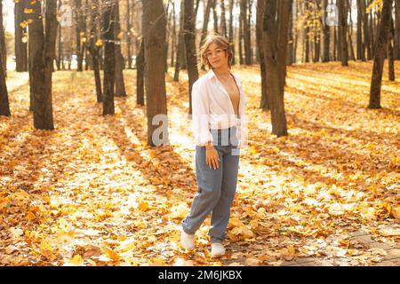 Portrait of young amazing teenage girl stand among trees on yellow fallen maple leaves in park on sunny day in autumn. Stock Photo