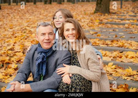 Portrait of family embracing, sitting on concrete stairs covered with yellow fallen leaves among trees in park forest. Stock Photo