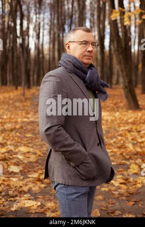Portrait of middle-aged man standing, putting hands into pockets near trees among yellow leaves in forest in autumn. Stock Photo