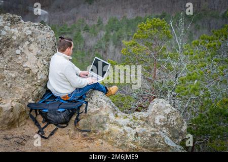 Travel man working with laptop sitting on the rocky mountain on beautiful scenic clif background Stock Photo