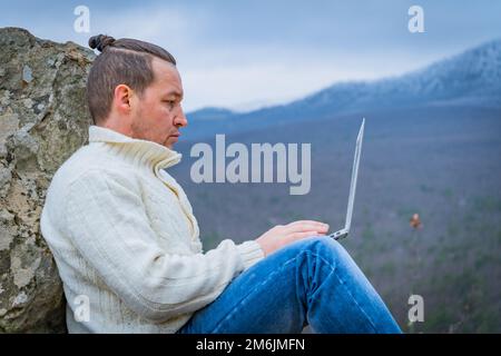 Hipster man working with laptop sitting on the rocky mountain on beautiful scenic clif background Stock Photo